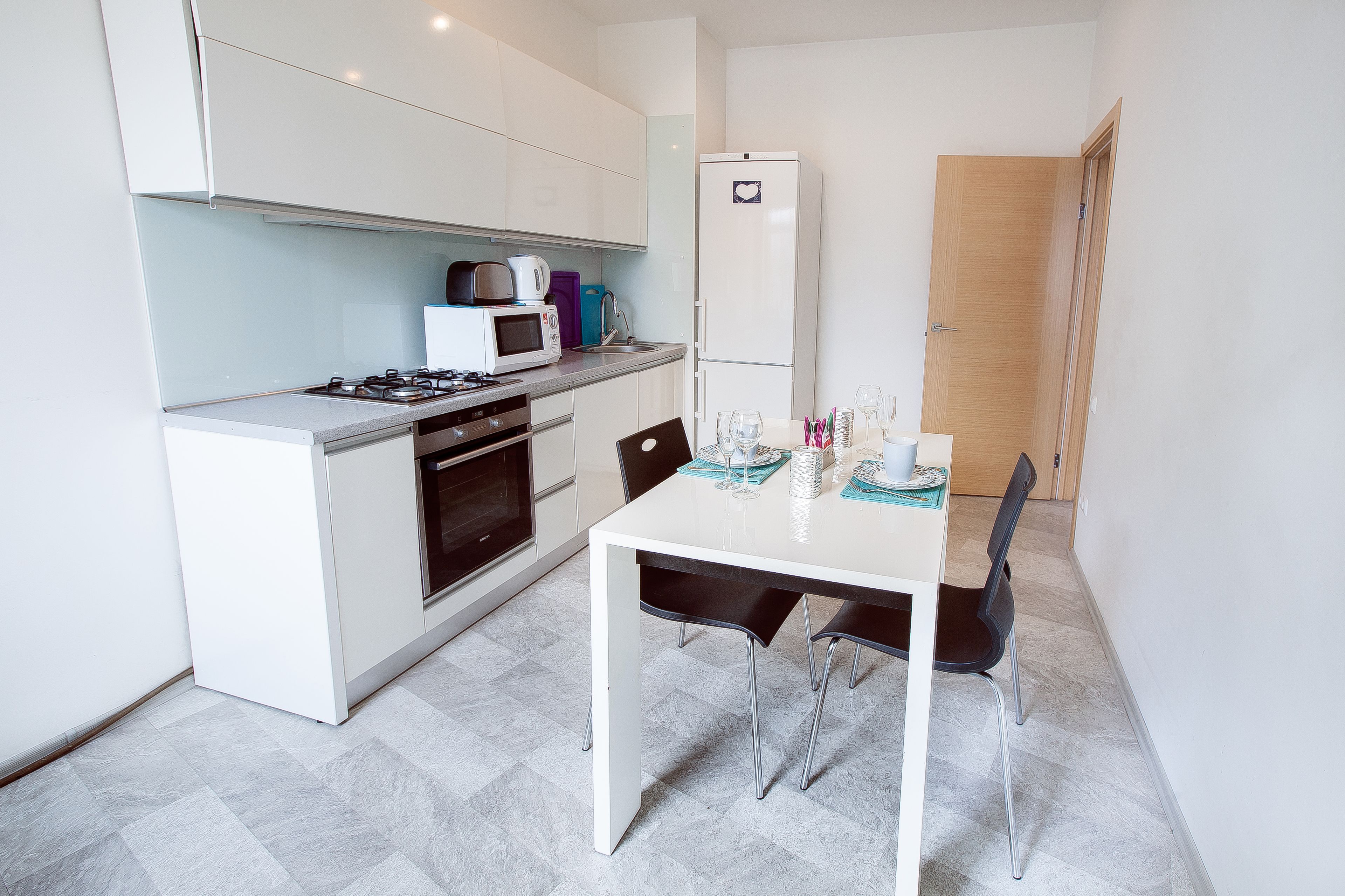 Une cuisine compacte avec des meubles blancs, une cuisinière, un four à micro-ondes et un réfrigérateur. On y trouve une table à manger avec deux chaises et un petit îlot central doté d’un évier.
