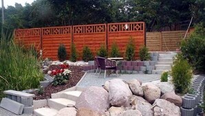 A garden with a wooden fence, a small patio with a table and chairs, and a variety of plants and rocks. - Hotel & Restaurant Kuhfelder Hof (Kuhfelde)