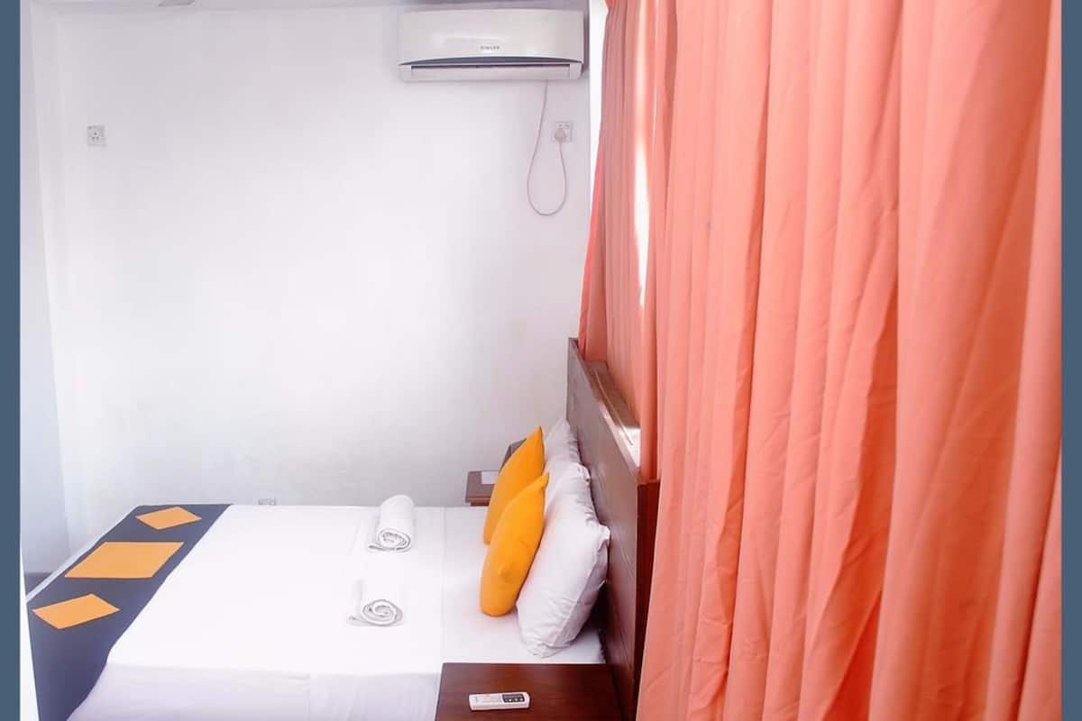 A small, neatly made bed with a white and orange pillow, a remote control on the side, and an air conditioning unit on the wall.