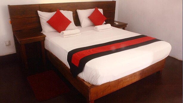Una cama bien hecha con sábanas blancas y detalles en rojo y negro, un cabecero de madera y una mesita de noche con una alfombra roja.