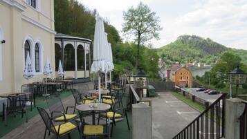 An outdoor seating area with tables and chairs, a covered umbrella, and a view of a river and hills in the background.