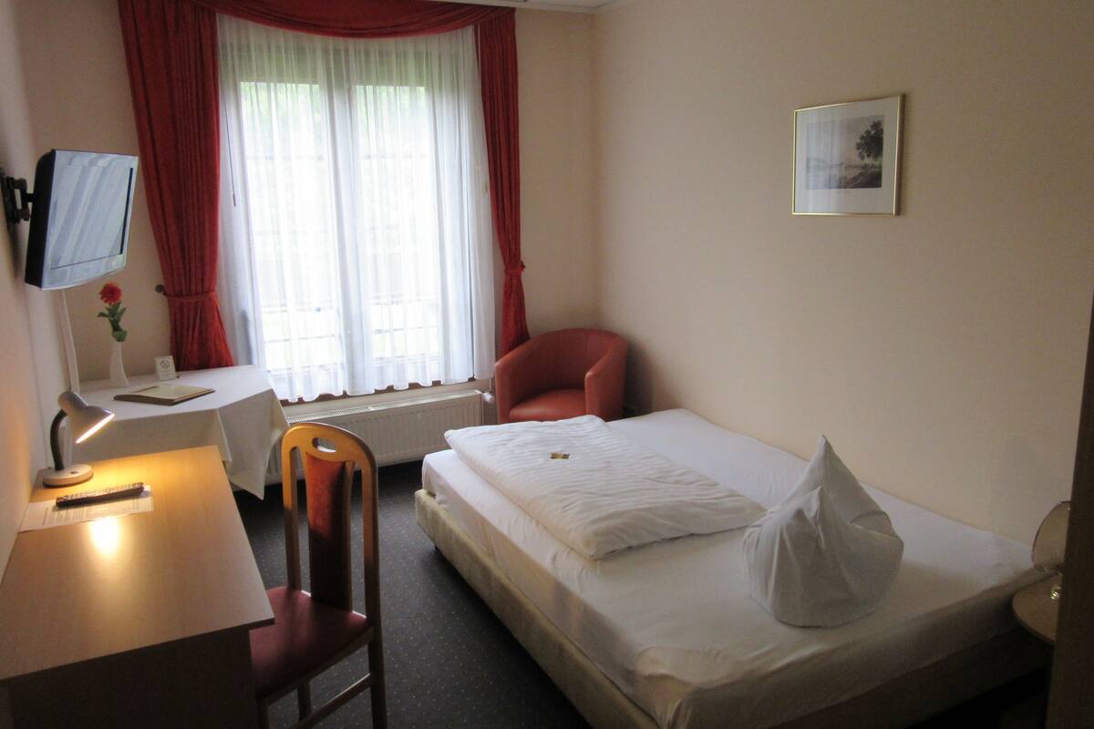 A hotel room with a single bed, a desk with a chair, a television, and a window with curtains.