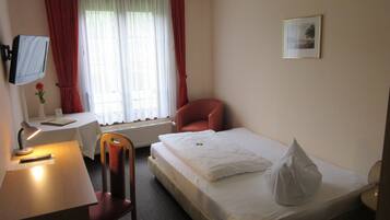 Ein Hotelzimmer mit einem Einzelbett, einem Schreibtisch mit Stuhl, einem Fernseher und einem Fenster mit Vorhängen.