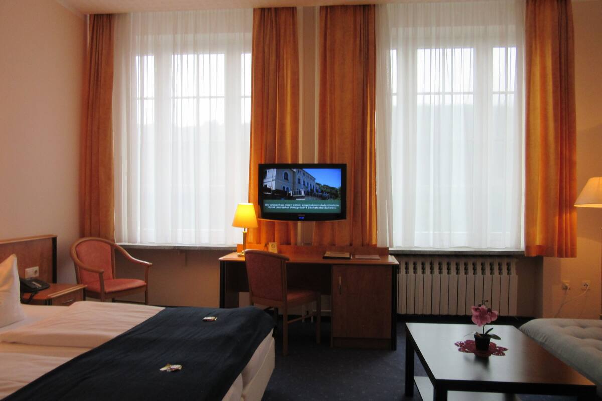 A hotel room with a bed, a desk, a television, a chair, and a table with a flower.