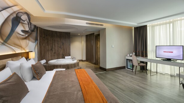 A modern hotel room with a bed, a desk with a TV displaying "WELCOME", a seating area with a table and chairs, and a large window with curtains. - B Business Hotel & Spa (Antalya)