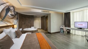 Habitación de hotel moderna con cama, escritorio con televisor mostrando “BIENVENIDOS”, zona de estar con mesa y sillas, y un ventanal con cortinas.
