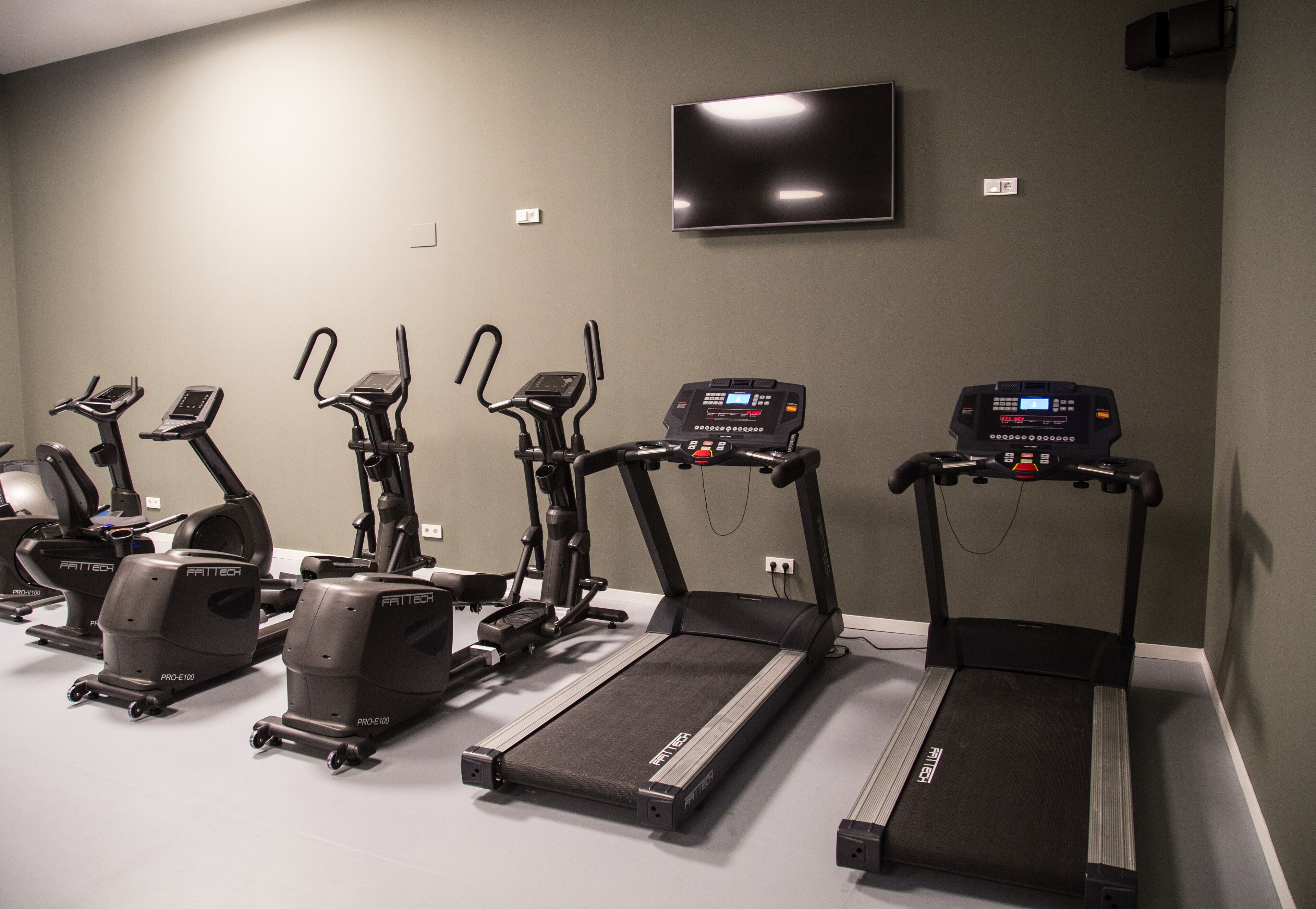 A fitness room with multiple cardio machines, including treadmills and elliptical trainers, arranged in a row.