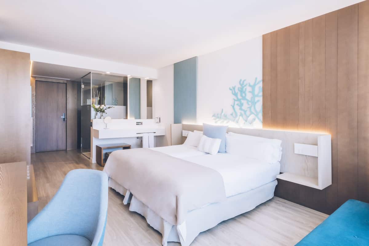 Ein modernes Hotelzimmer mit einem großen Bett, einem Schreibtisch, einem Sessel, einem Badezimmer mit Spiegel und einem an der Wand befestigten Regal.