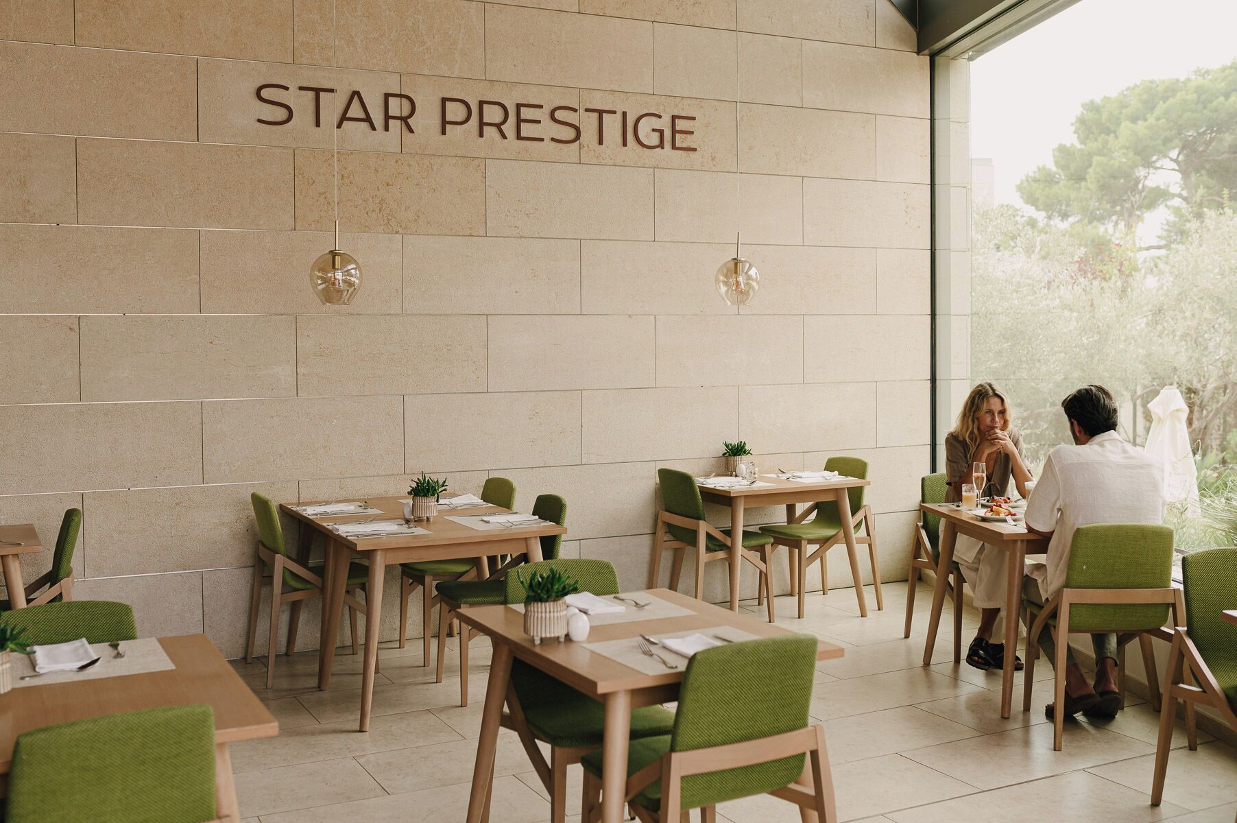 A restaurant interior with a sign reading "STAR PRESTIGE" on the wall, multiple tables with green chairs, and a view of greenery outside.