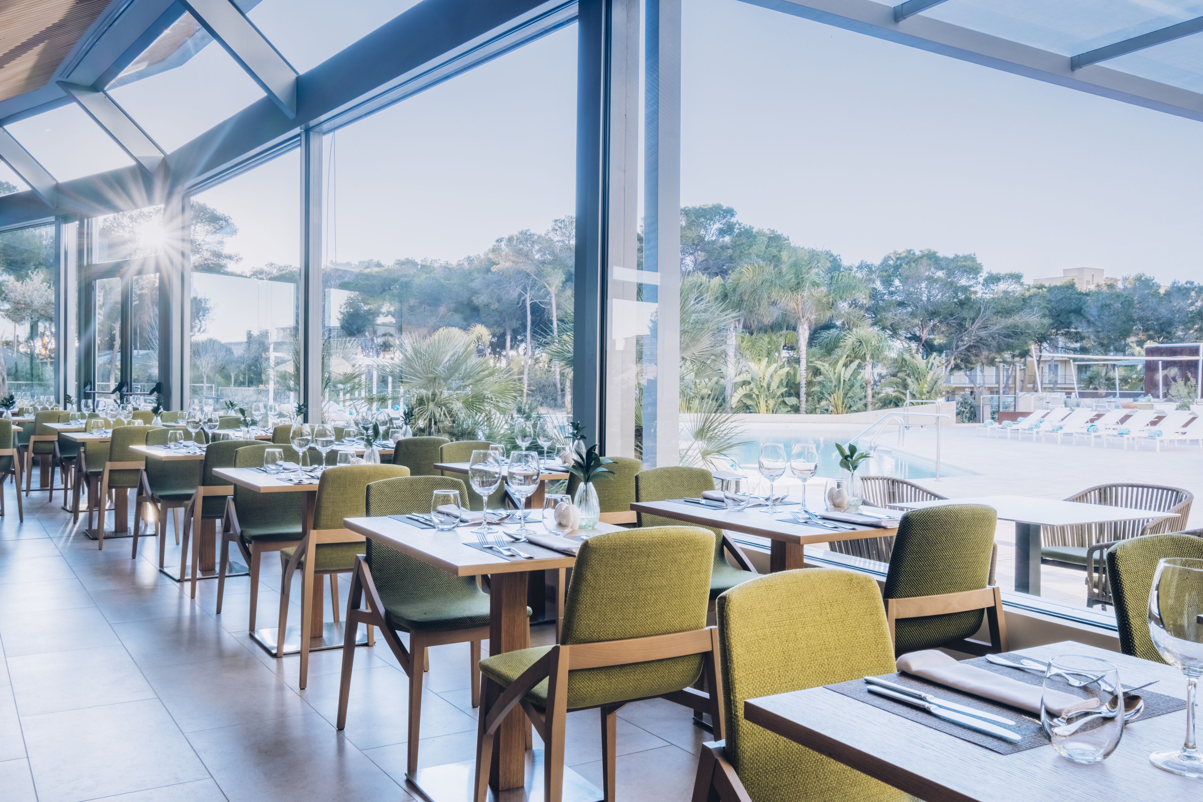 Un restaurante con mesas y sillas dispuestas para comer, con ventanas amplias que ofrecen vistas a una piscina y follaje.