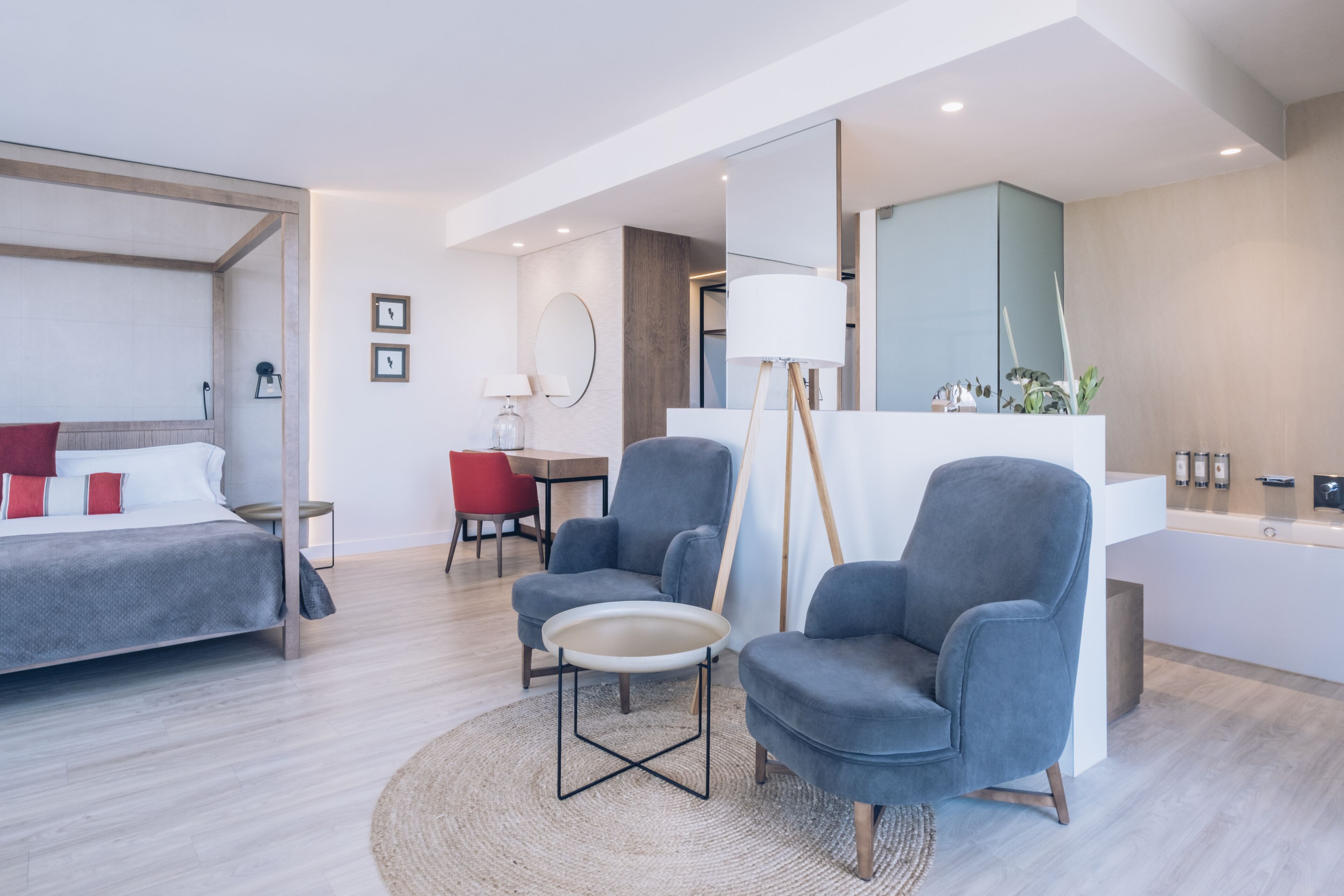 A modern hotel room with a bed, two armchairs, a small table, a desk, and a bathroom with a mirror and sink.