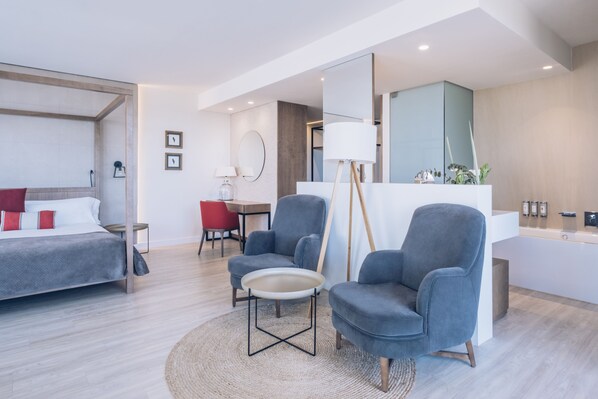 A modern hotel room with a bed, two armchairs, a small table, a desk, and a bathroom with a mirror and sink.