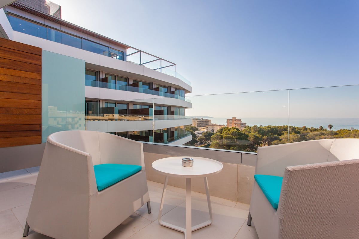 Ein Balkon mit zwei weißen Korbstühlen und einem kleinen weißen Tisch, mit Blick auf ein modernes Gebäude und das Meer.