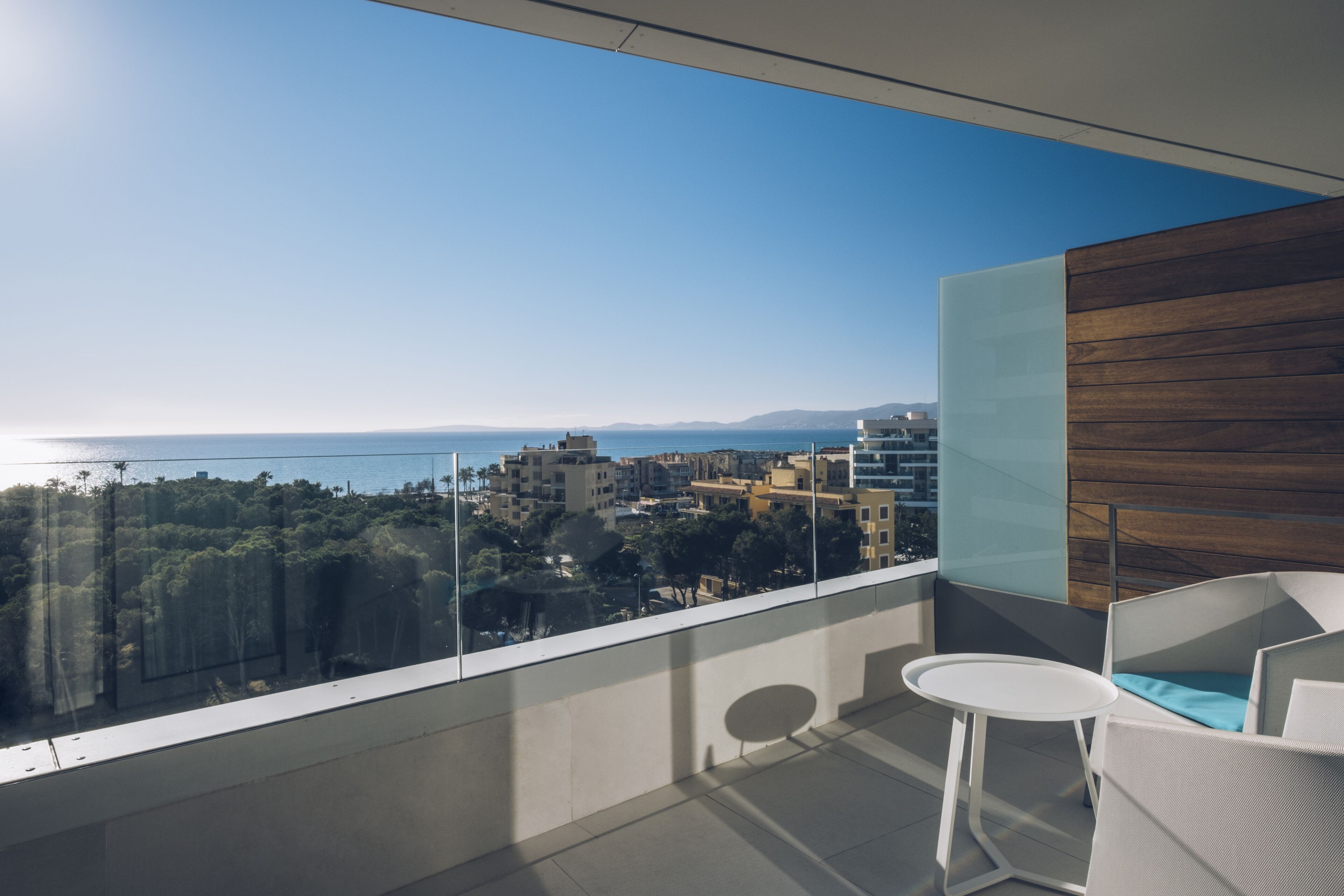 Un balcón con vista al mar y edificios, una silla blanca y una pequeña mesa redonda.