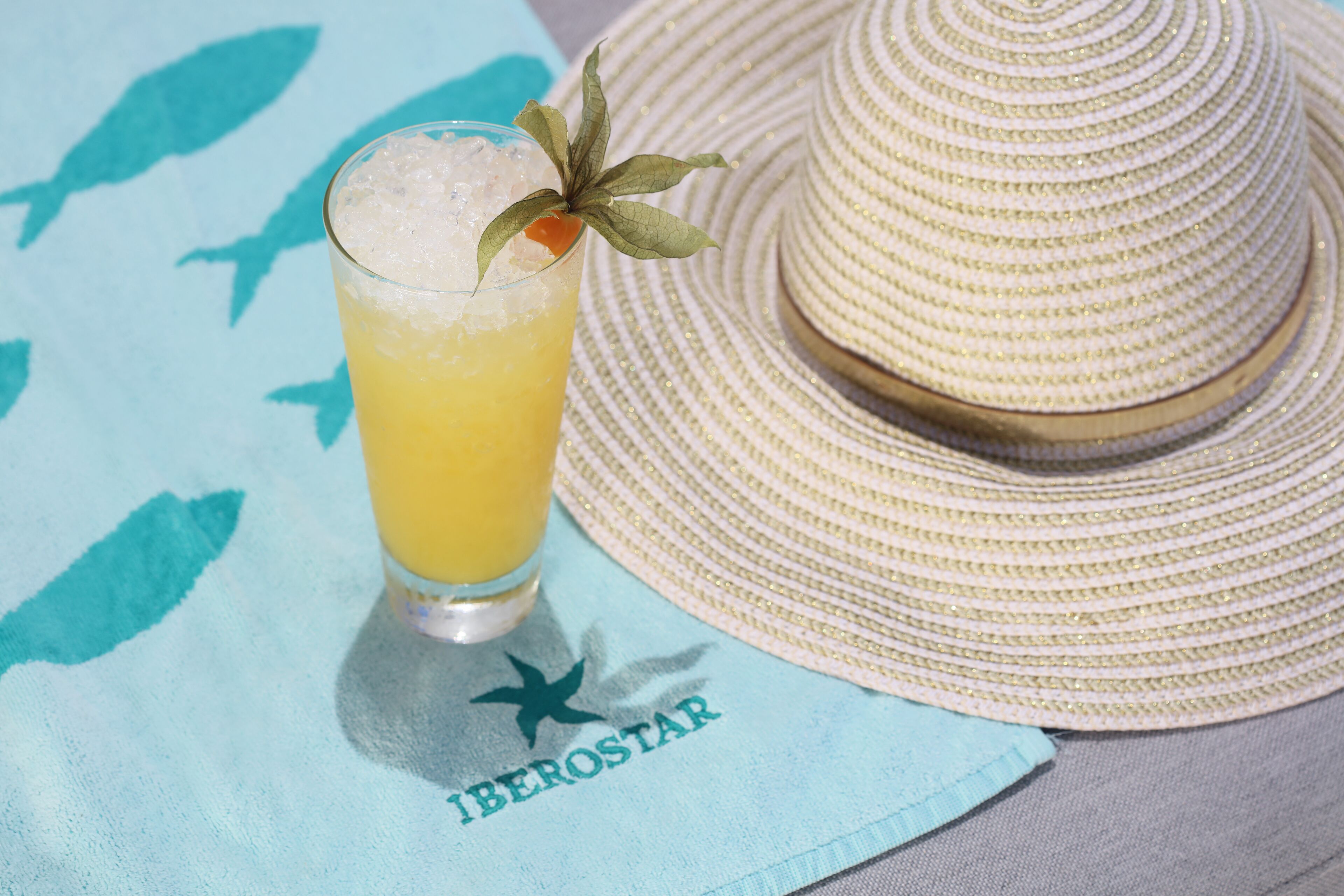 Un sombrero de paja, un vaso de una bebida amarilla con hielo y una ramita de decoración, y una toalla con una estrella y la palabra 'HEROSTAR'.
