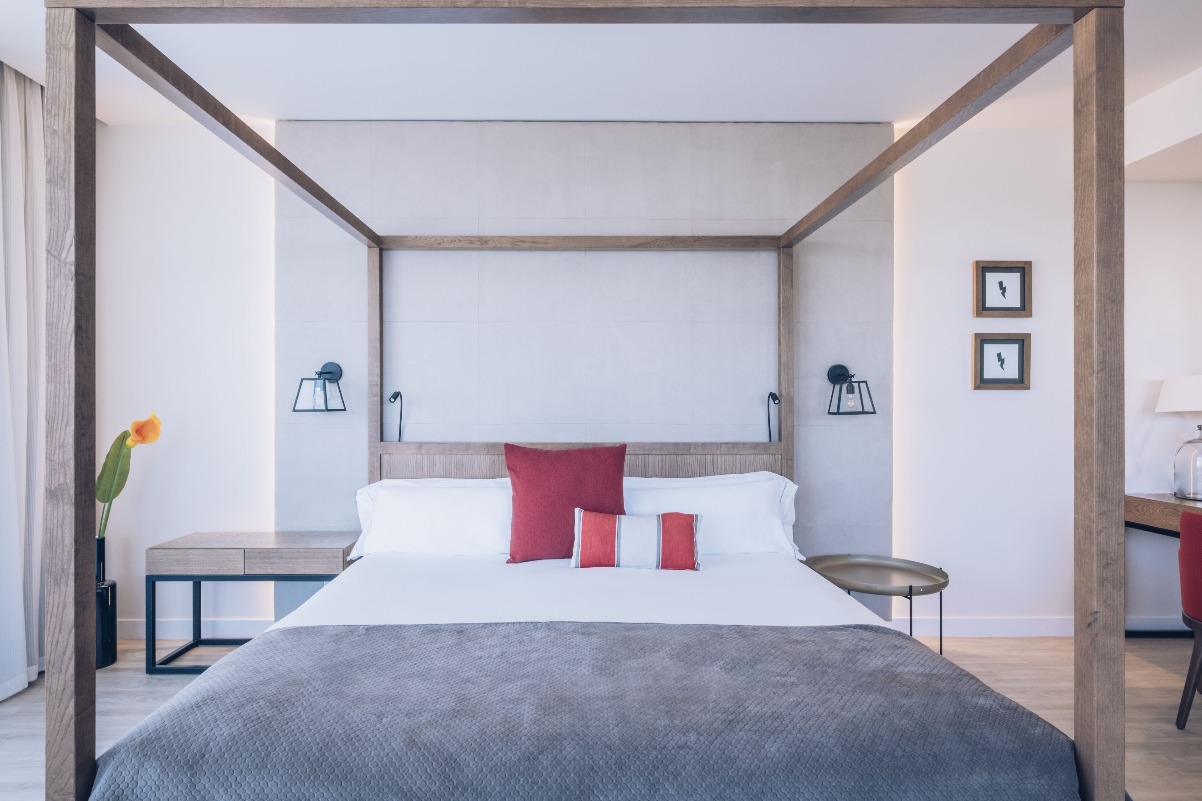 A modern hotel room with a four-poster bed, a bedside table, a lamp, and a small desk with a chair.