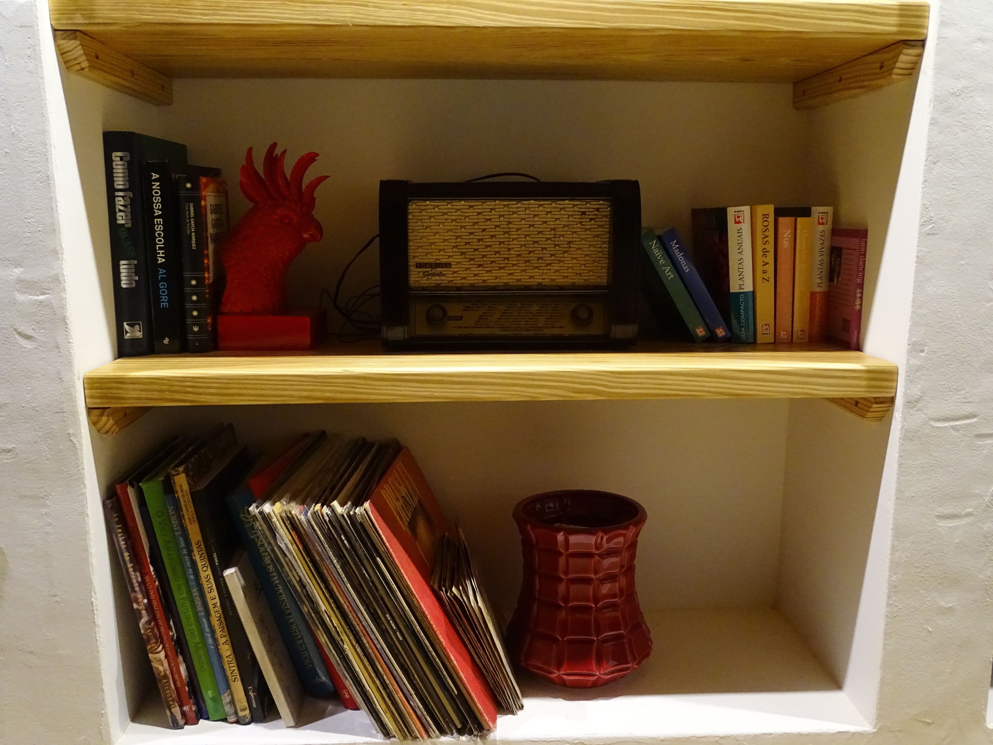 Uma estante com livros, um objeto decorativo vermelho e um rádio antigo.