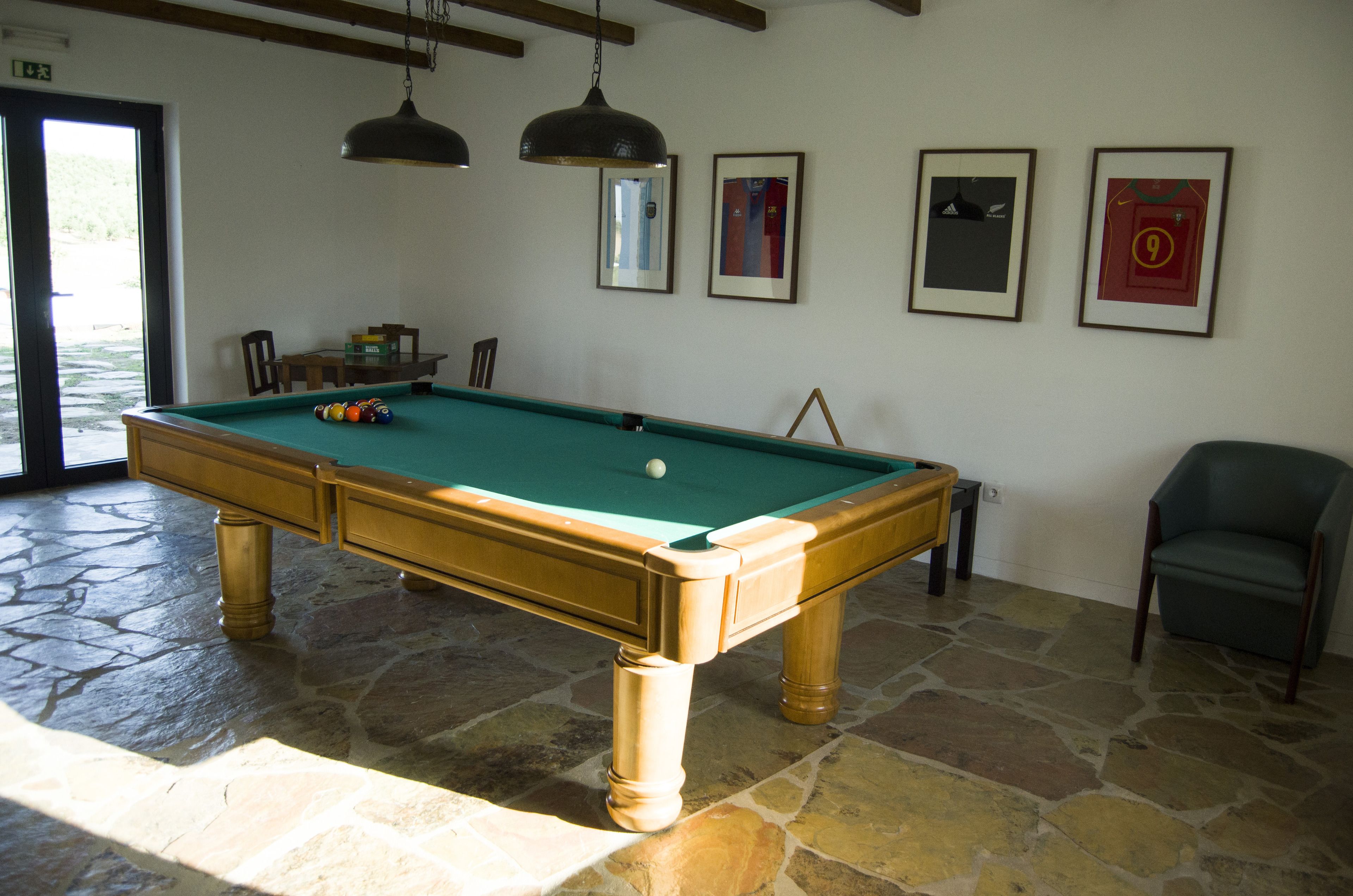 A pool table in a room with stone flooring, wooden furniture, and framed sports jerseys on the wall.