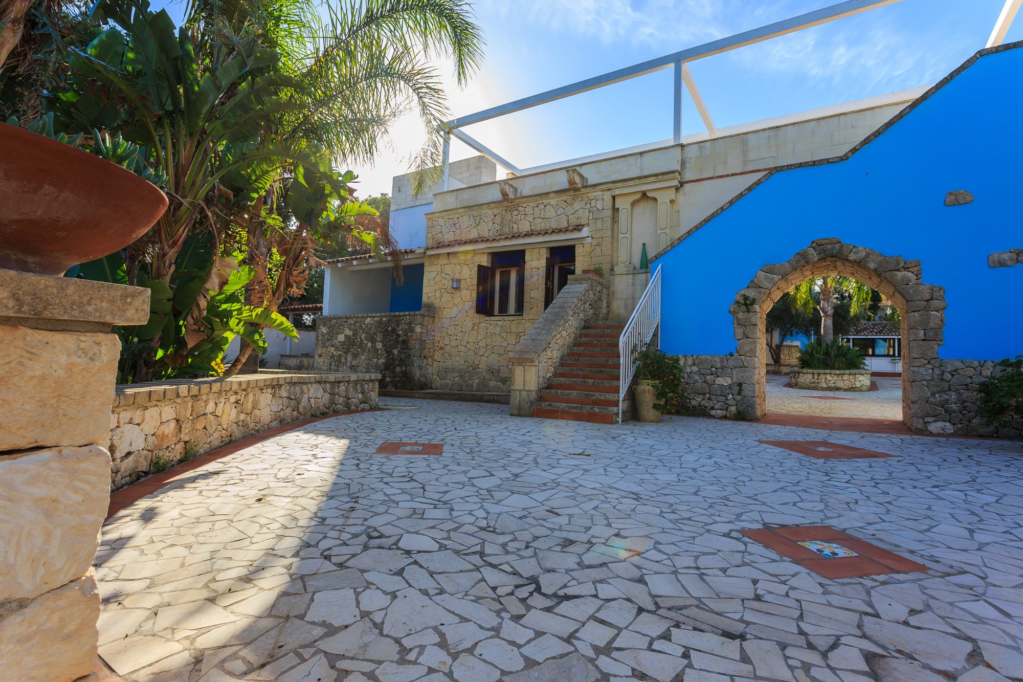 A courtyard with stone flooring, a blue building with a stone archway, and a staircase leading to a door.