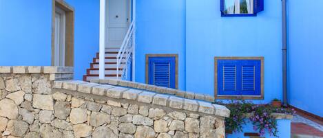 A blue building with a stone wall in front, a white door, and a staircase leading to a second floor.