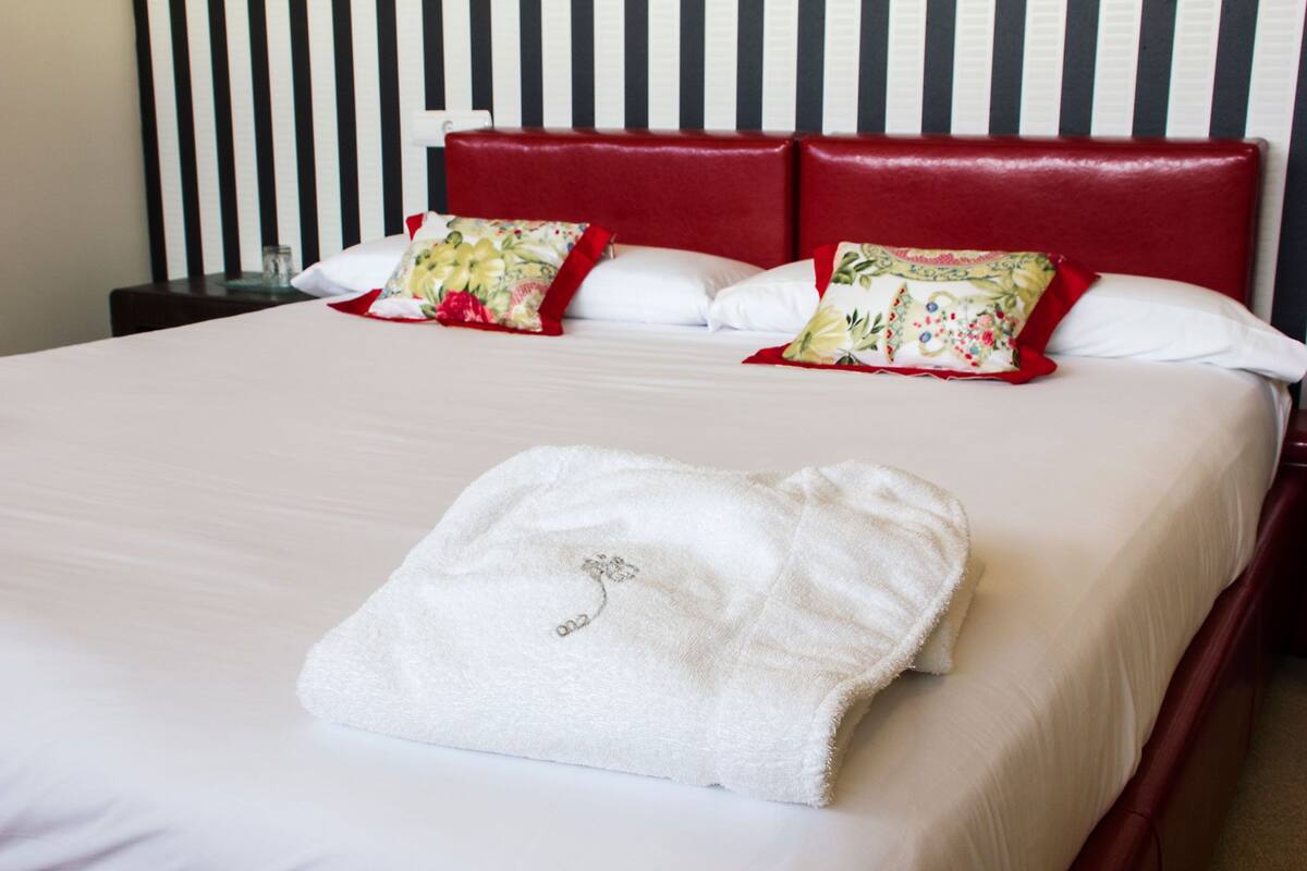 Una cama bien hecha con edredón blanco, dos cabeceras rojas y almohadas con motivos florales.