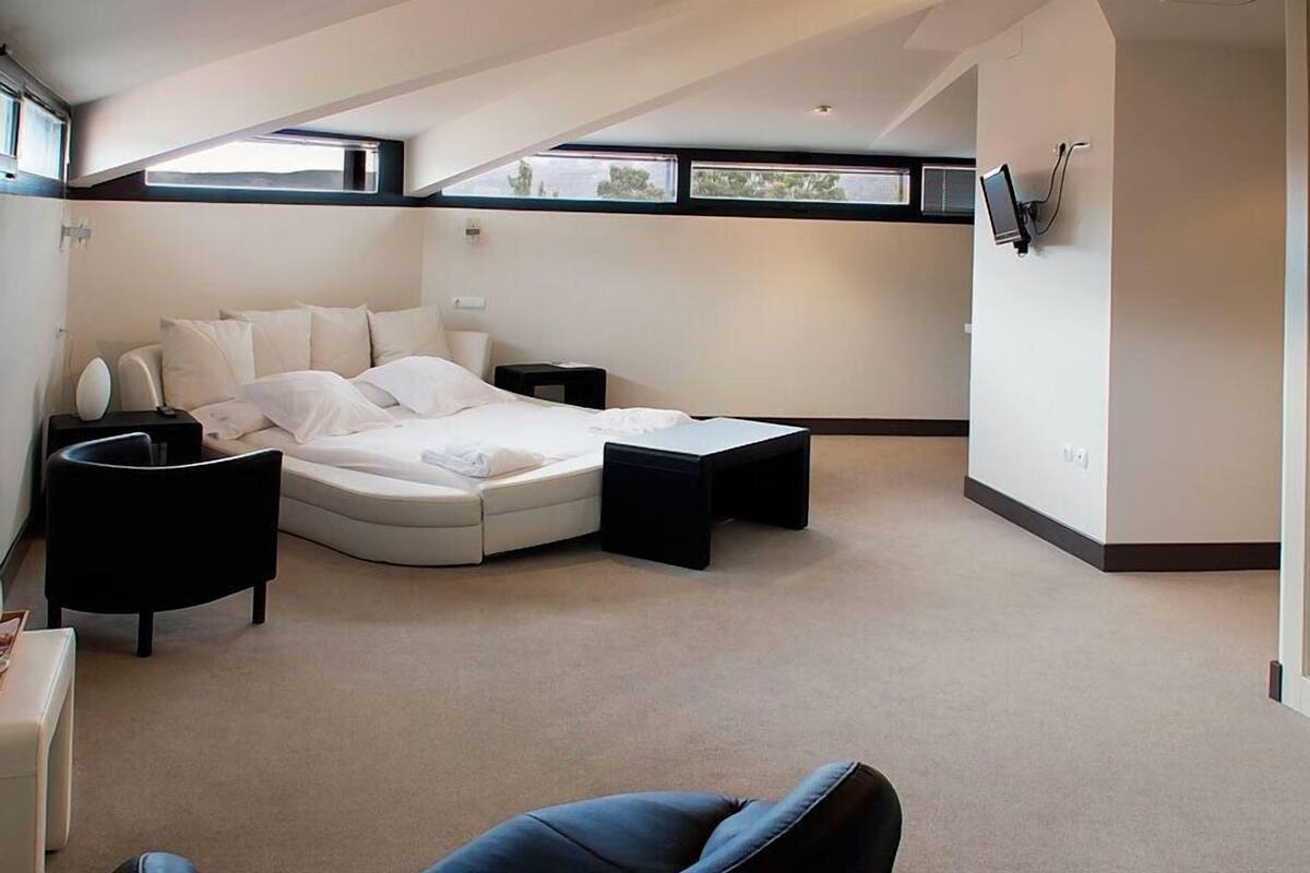 Un dormitorio moderno con una cama curvada, un reposapiés negro, una silla negra y una mesita de noche blanca.