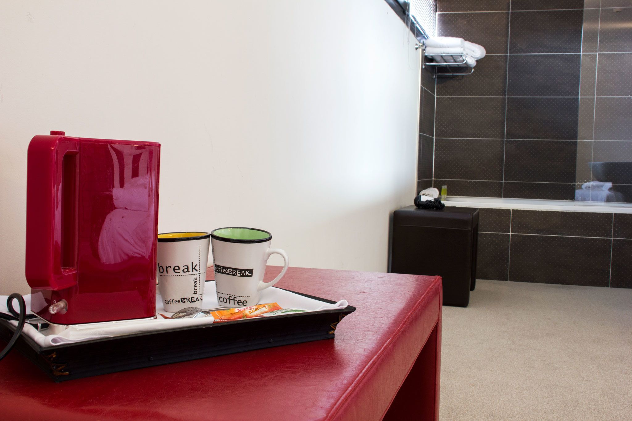 A red coffee maker, two coffee mugs, and a tray with coffee packets on a table in a bathroom with dark tiled walls.