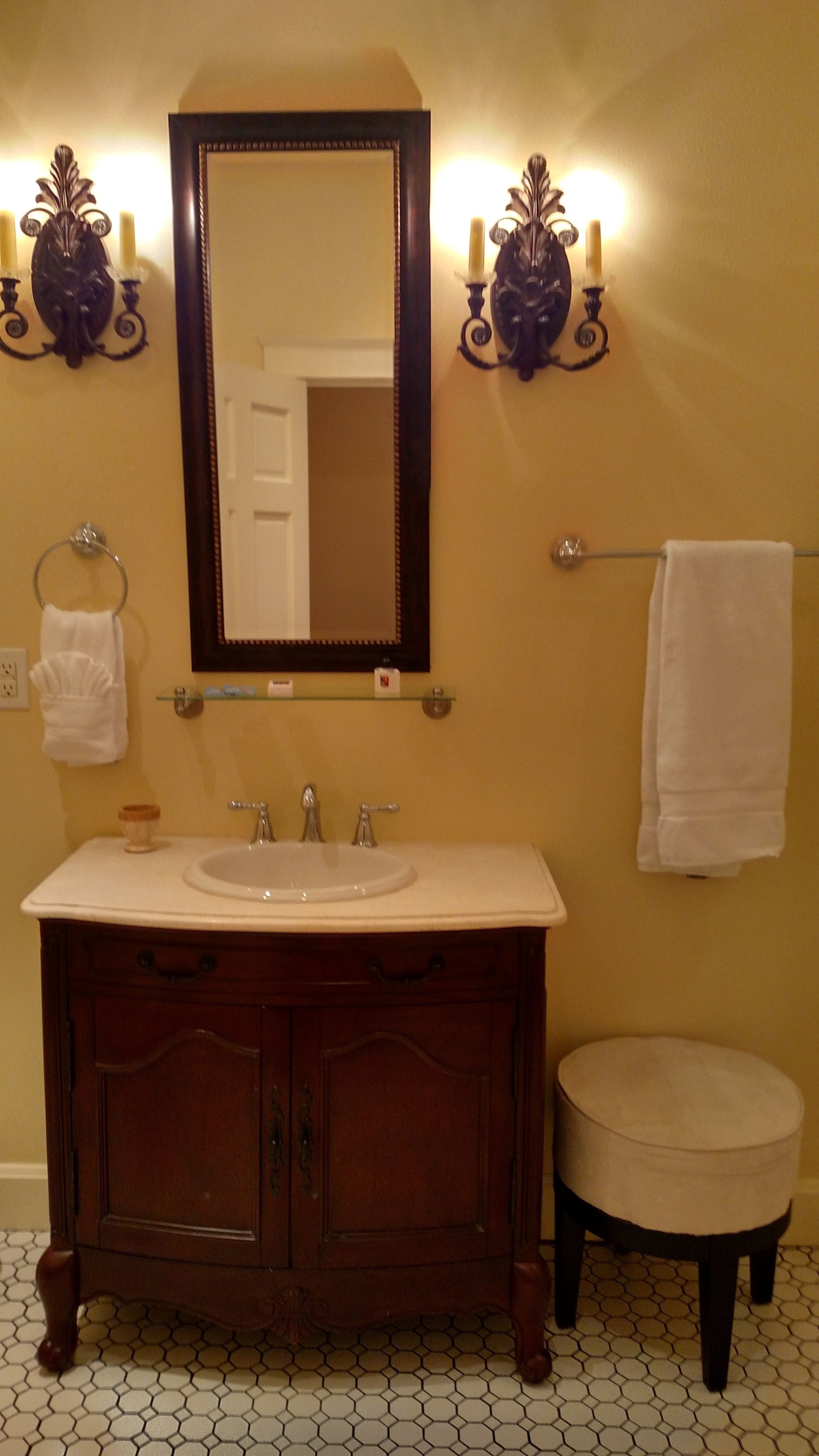 Une salle de bain avec un meuble-lavabo en bois, un grand miroir, deux vasques, un porte-serviettes et un tabouret rond.