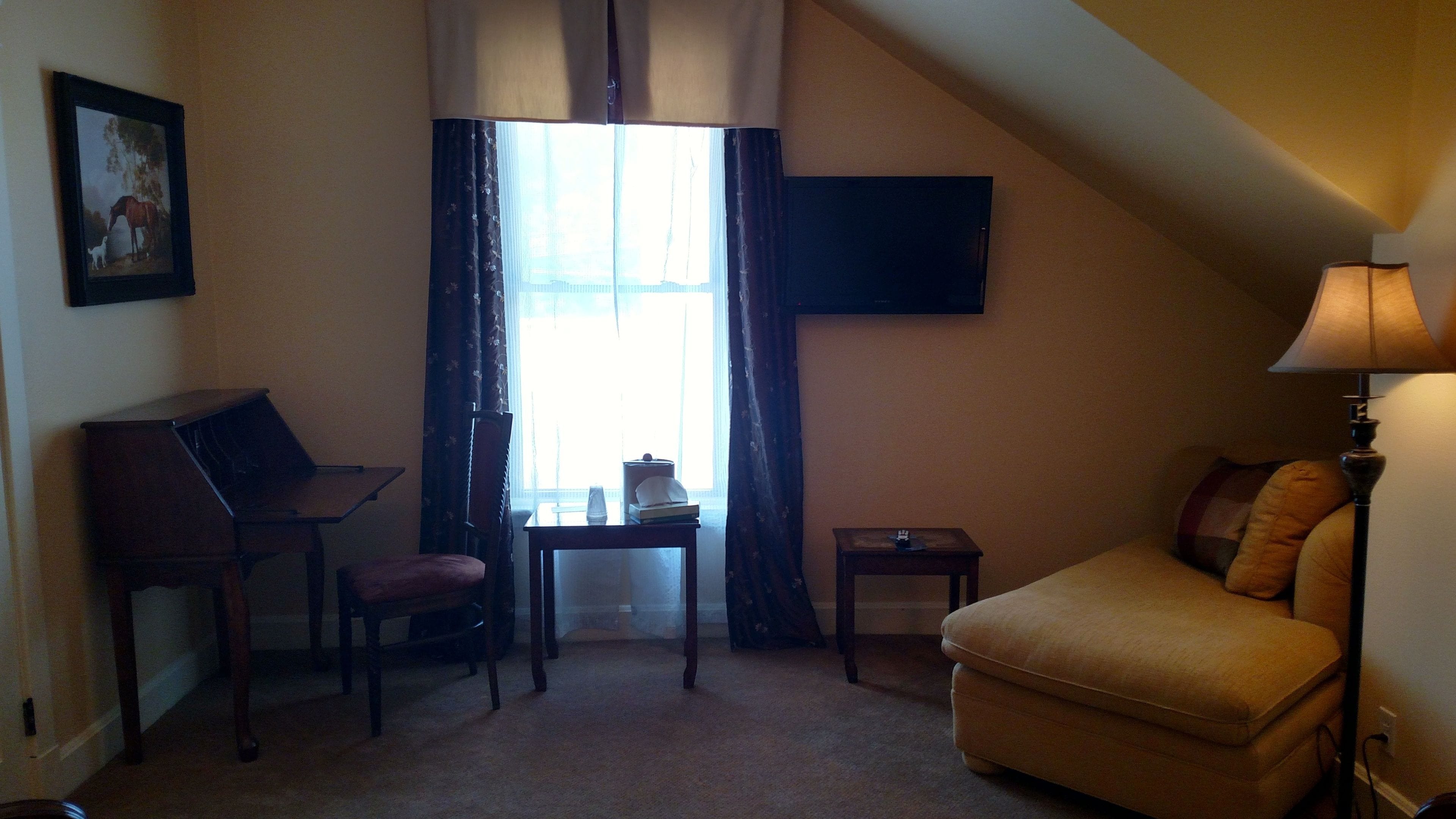 A small, cozy room with a desk, chair, and a sofa. There is a window with curtains, a flat-screen TV mounted on the wall, and a lamp on a side table.