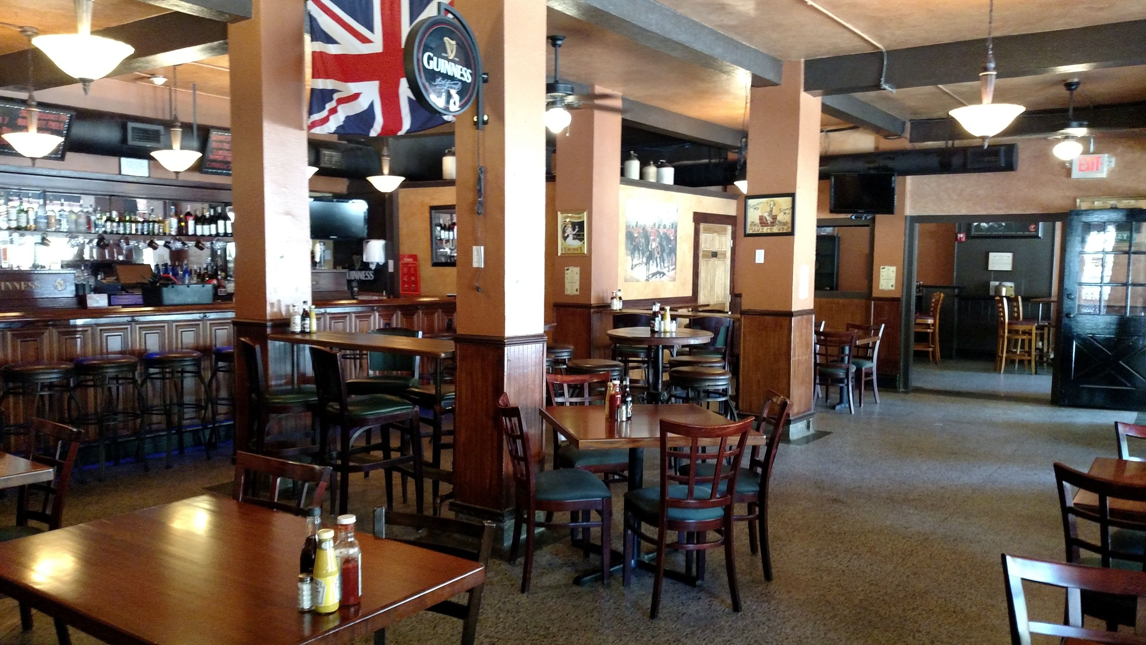 Interior de restaurante con mesas y sillas de madera, una zona de bar con botellas y una gran bandera del Reino Unido.