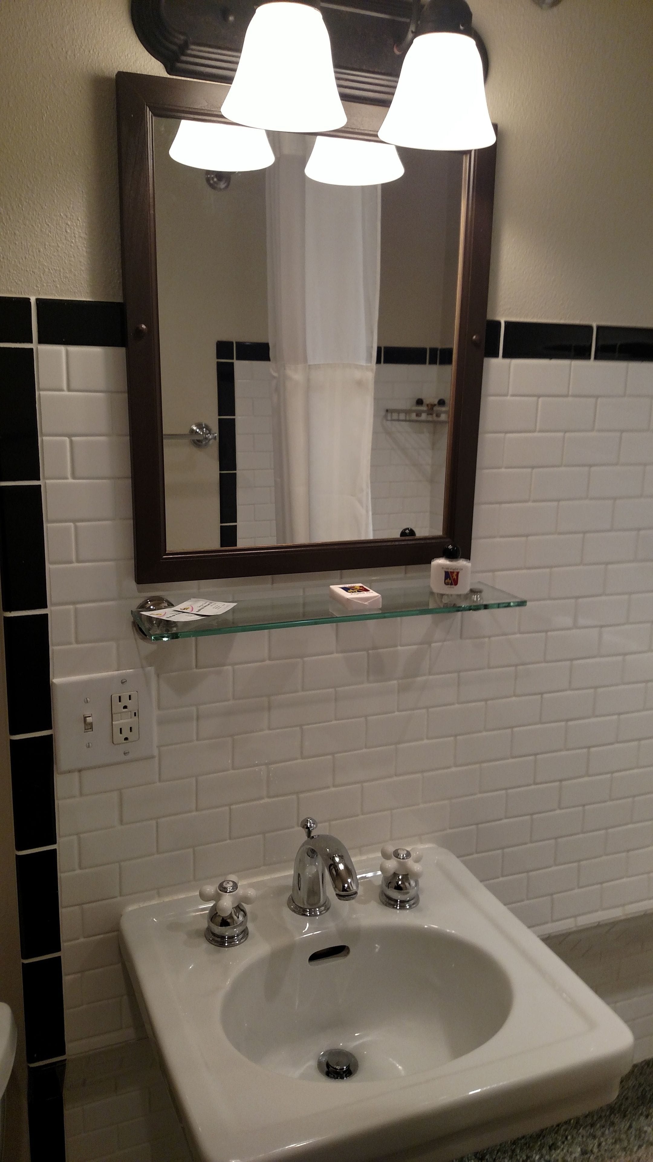 Un baño con un lavamanos blanco, tres grifos, un espejo con marco de madera y un estante de vidrio sobre el lavamanos.