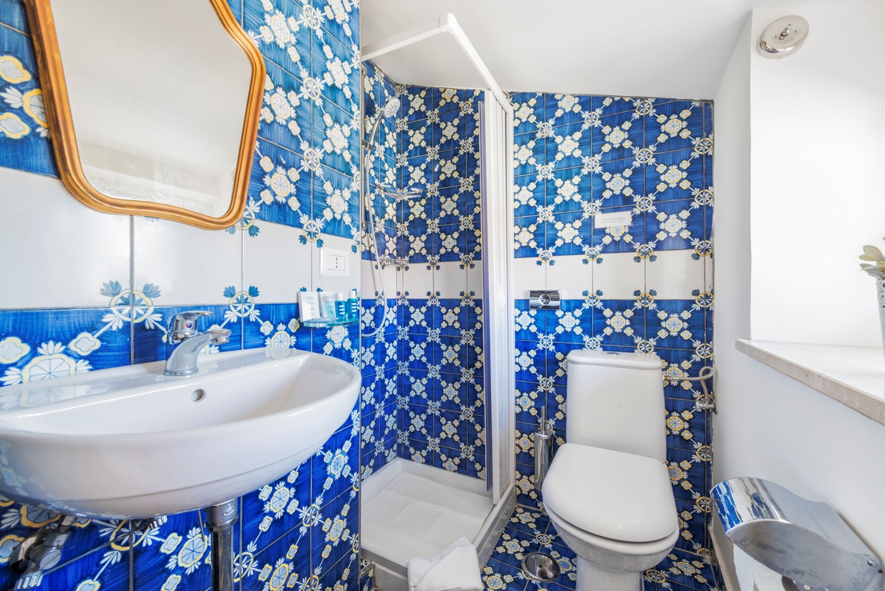 Un baño con lavamanos, inodoro y ducha blancos. Las paredes están cubiertas de azulejos con motivos florales azules y blancos.