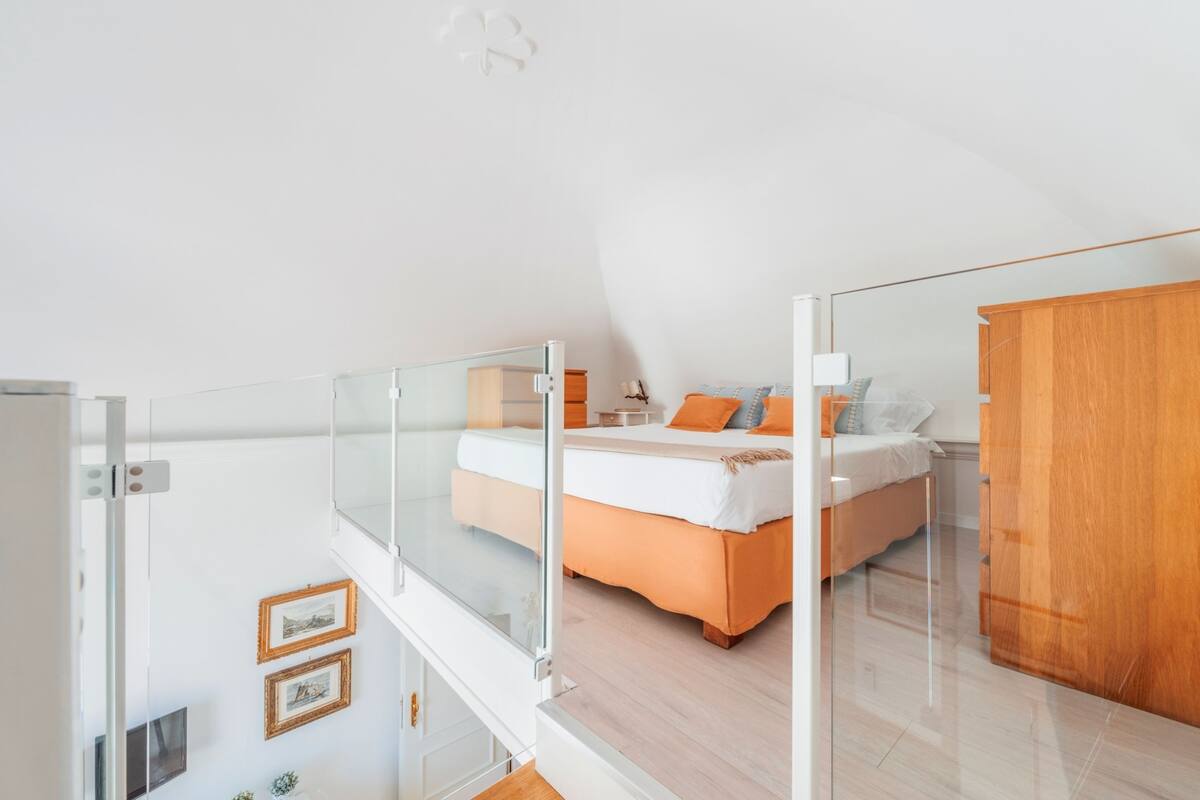 A modern bedroom with a glass balcony, a bed with orange bedding, and a wooden wardrobe.