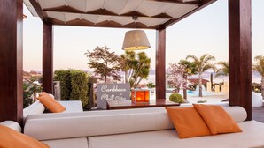 A lounge area with a sign reading "Caribbean Chill Spa," a white sofa with orange cushions, a hanging lamp, and a view of palm trees and a pool.