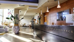 A hotel reception area with a modern design, featuring a reception desk labeled "GUEST EXPERIENCE," a seating area with sofas, and a large potted plant.