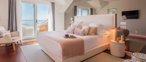 A modern hotel bedroom with a large bed, a balcony with a view of the sea, and a flat-screen TV.