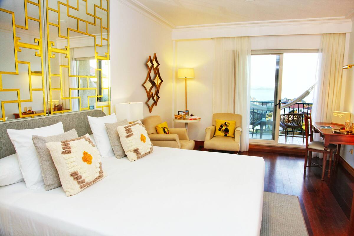 Quarto de hotel com uma cama grande, duas poltronas, uma escrivaninha e uma varanda com vista.