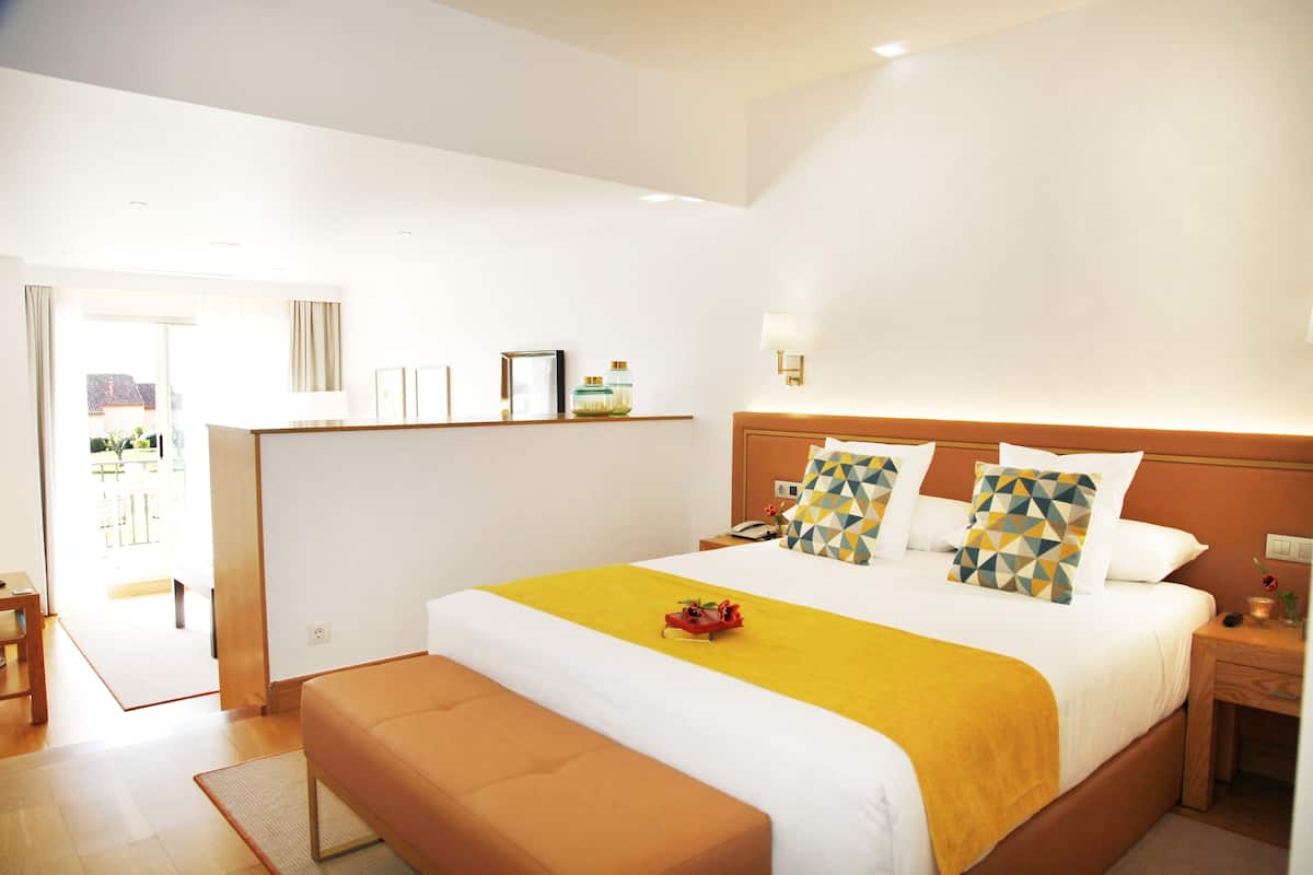 Quarto de hotel com uma cama grande, uma mesinha de cabeceira, um banquinho e vista para o exterior.