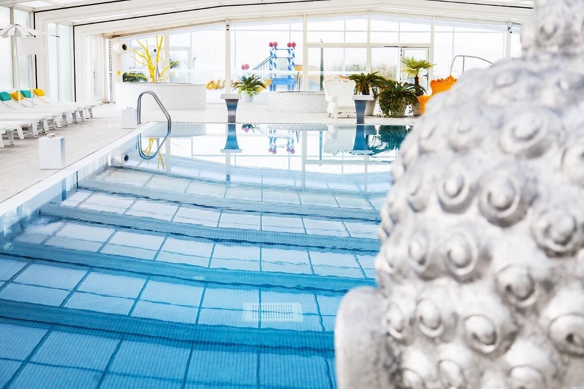 Piscina interior con agua cristalina, un trampolín y una zona de estar con sillones reclinables y plantas en macetas.