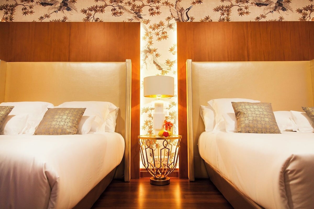 Quarto de hotel com duas camas, uma mesa de cabeceira central com abajur e papel de parede floral.