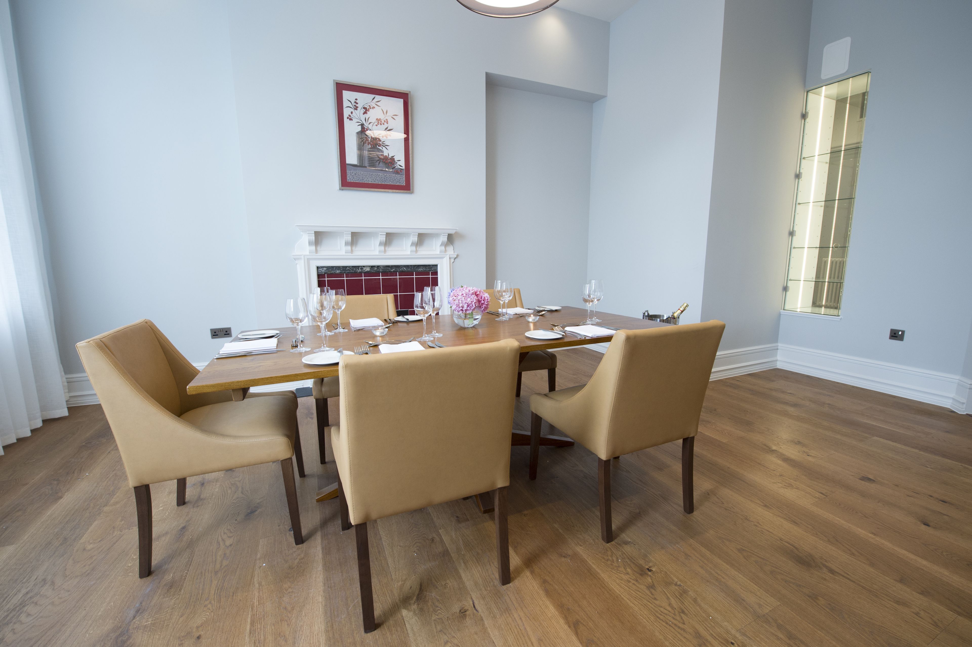 A dining room with a wooden table set for four, beige chairs, a white fireplace, and a framed picture on the wall.
