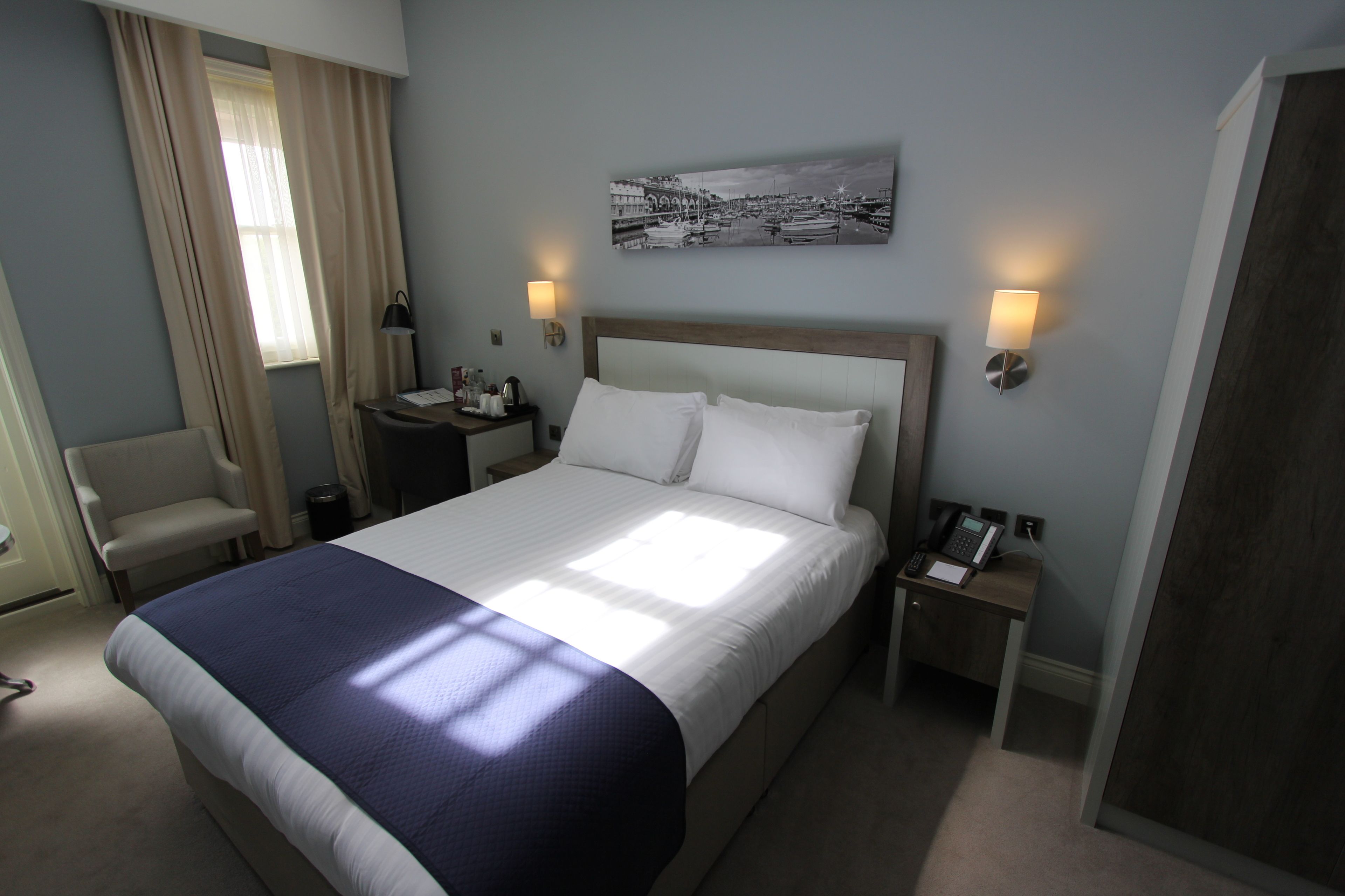 A hotel room with a large bed, a chair, a desk with a telephone, and a wall-mounted lamp.