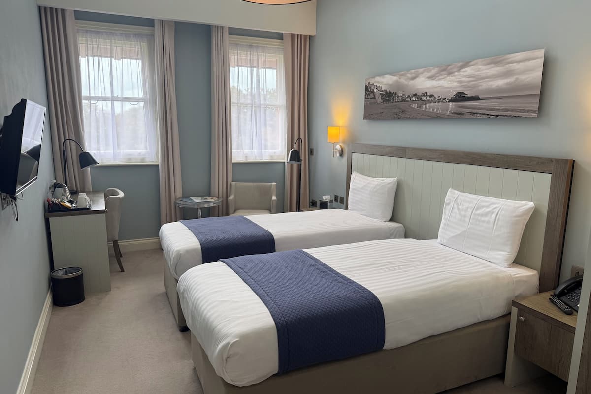 A hotel room with two beds, a desk, a chair, a TV, a lamp, and a painting on the wall.