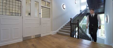 A spacious room with a wooden floor, a staircase with a black railing, and large windows with a grid pattern.
