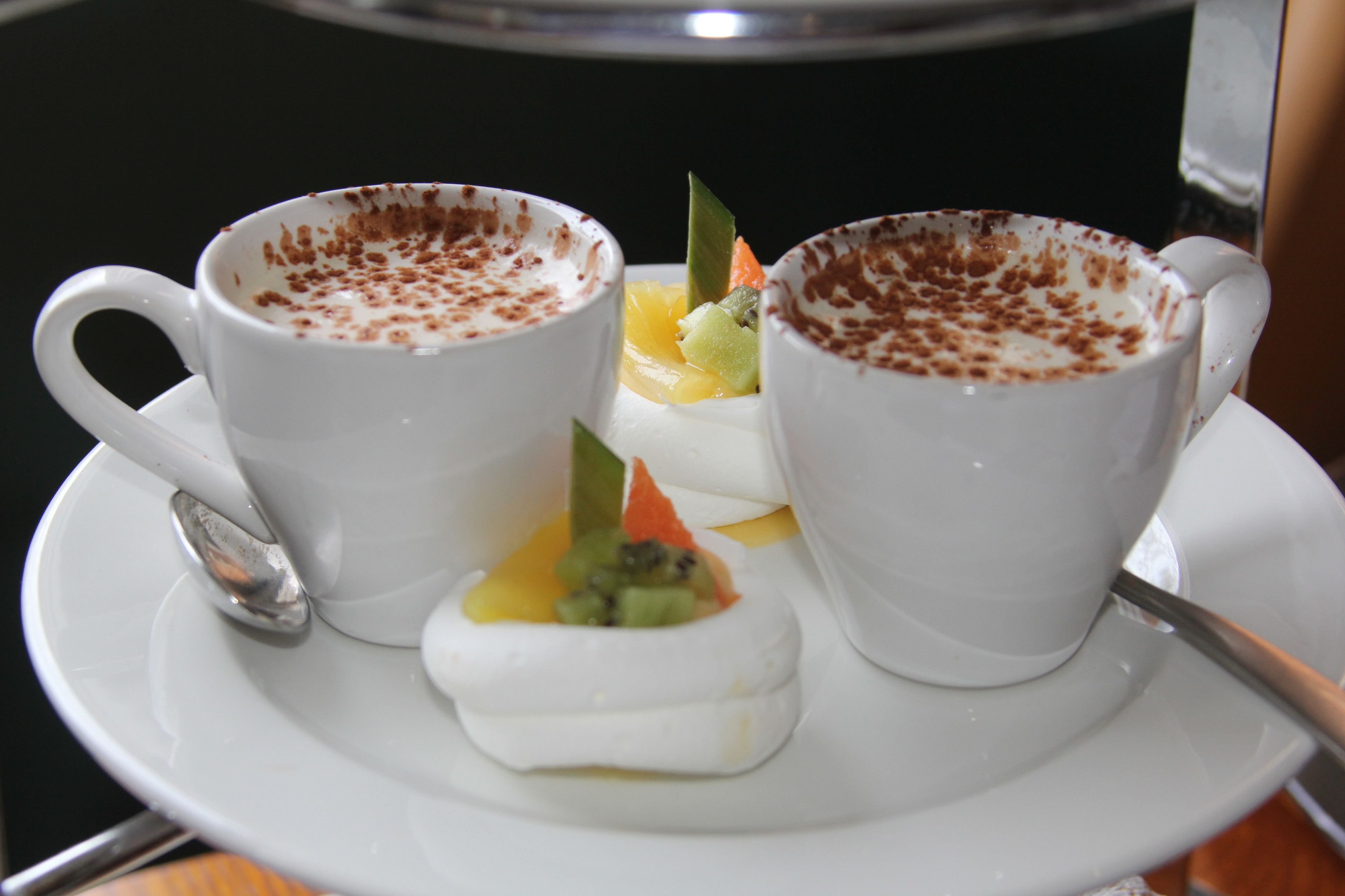 Two white cups with a frothy top, a small plate with fruit, and a spoon on a saucer.