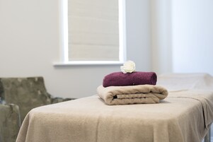 Cama arrumada com um cobertor bege e duas toalhas dobradas, uma roxa e outra bege, com uma flor branca em cima.