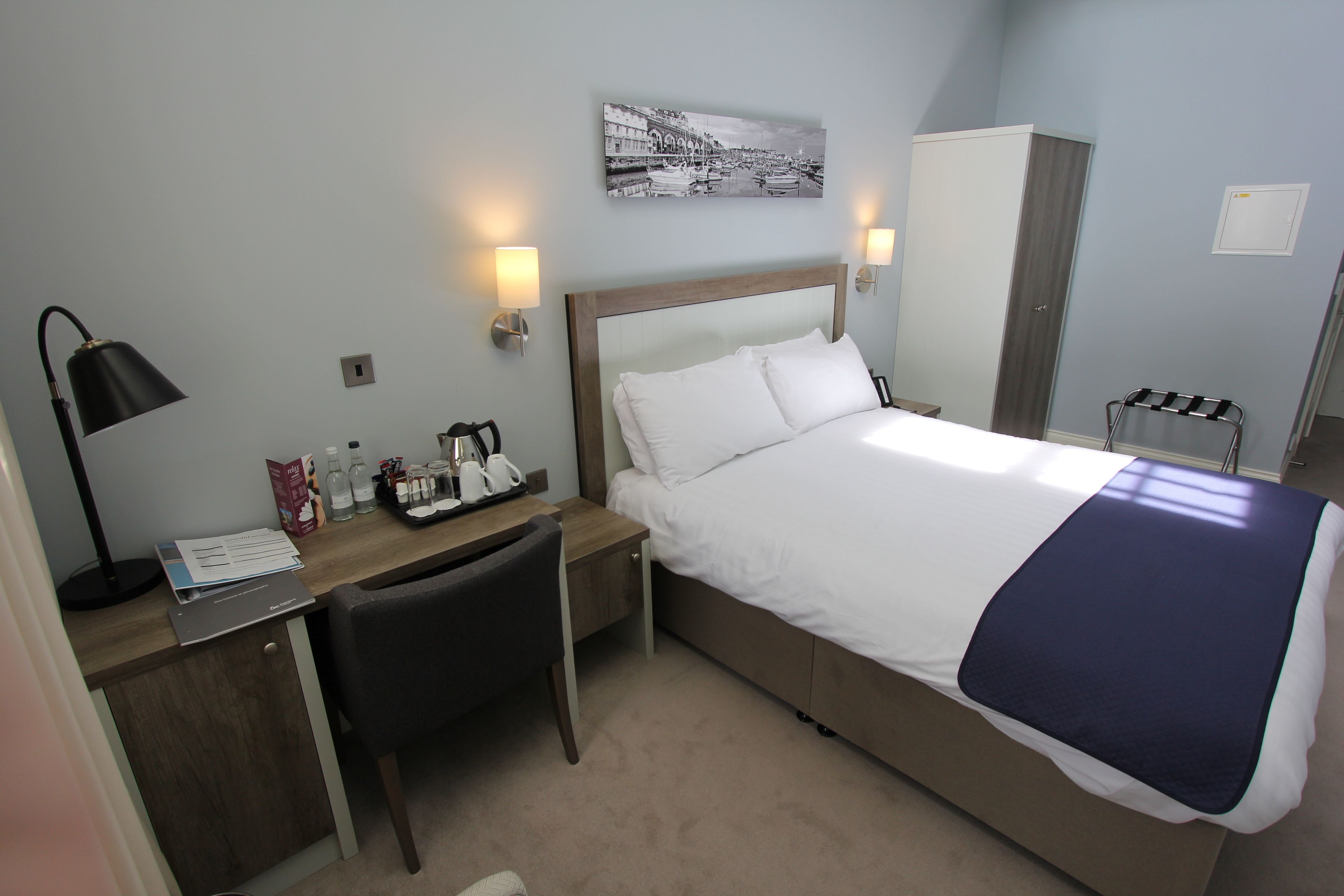 Habitación de hotel con cama, mesita de noche, lámpara, silla, escritorio y un cuadro colgado en la pared.