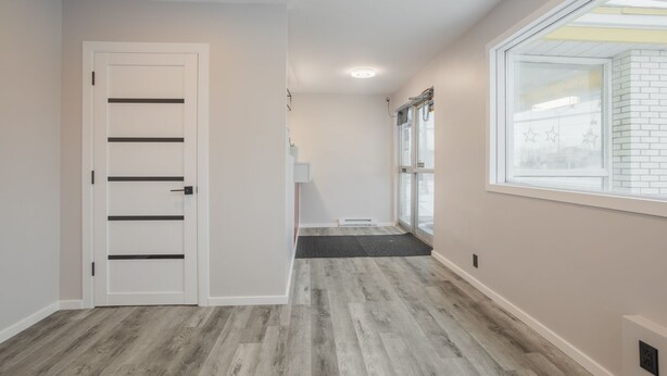 Un couloir avec une porte blanche, une fenêtre donnant sur un bâtiment en briques et un plancher en bois gris.