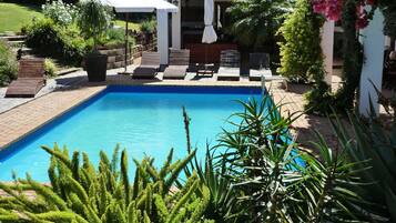 A rectangular swimming pool with a tiled deck, surrounded by lounge chairs and a pergola with umbrellas.