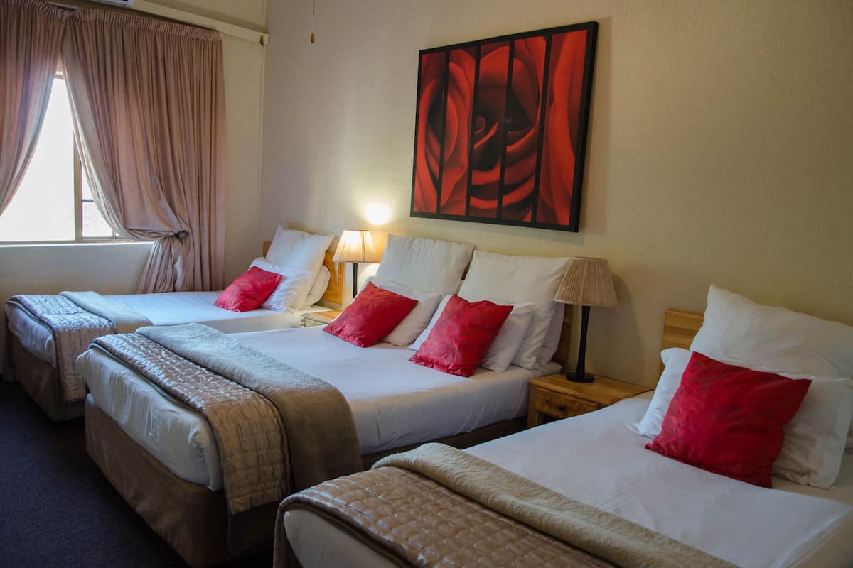 A hotel room with three single beds, each with white bedding and red pillows. There is a painting on the wall above the beds, a window with curtains, and a lamp on each bedside table.